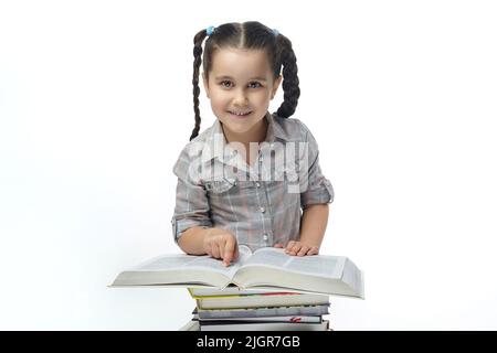 charmante petite fille avec des picots tressés se trouve devant un grand livre et regarde dans l'appareil photo. Banque D'Images