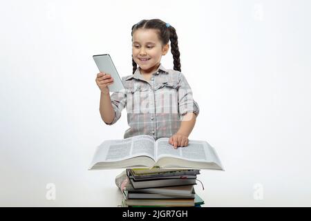 charmante petite fille avec des picots tressés est assise devant un grand livre et regardant l'écran de son smartphone. Banque D'Images