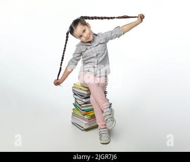 charmante petite fille avec des picots tressés est assise sur une grande pile de livres. Banque D'Images