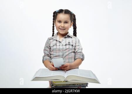 charmante petite fille est assise devant une grande pile de livres et tient un smartphone dans ses mains. Banque D'Images