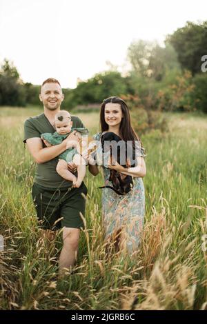 Beaux et heureux parents caucasiens jouant avec leur fille de six mois et leur chien marchant sur le terrain vert. Famille positive de trois personnes profitant de temps libre sur l'air frais. Banque D'Images