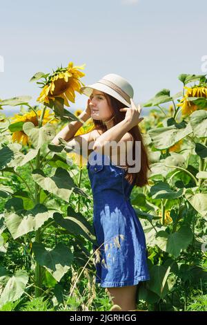 Une belle fille dans une robe bleue et un chapeau blanc d'été se tient dans un champ de tournesols près d'une grande fleur Banque D'Images