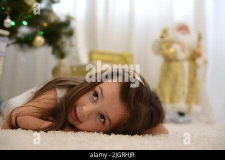 La fille se trouve sous l'arbre et regarde vers la gauche au lieu du Père Noël dans un jouet en costume doré et elle se trouve sur un tapis blanc. Photo de haute qualité Banque D'Images