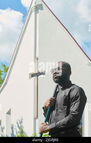 Jeune pasteur sérieux de l'église luthérienne en lunettes et chemise noire avec collier de bureau tenant le rake contre le bâtiment blanc avec la croix Banque D'Images
