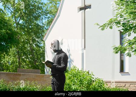 Vue latérale du jeune prédicateur en vêtement noir regardant dans la Bible sainte ouverte tout en lisant les versets de l'un des Canonic Gosels dehors Banque D'Images
