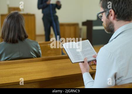 Homme d'âge moyen avec la Sainte Bible ouverte lisant les versets de l'Evangile tout en étant assis sur le banc par bureau et en écoutant le sermon pendant le service de l'église Banque D'Images