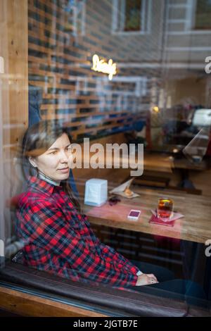 Une femme dans un café attend sa date, regardant par la fenêtre, il y a du thé et du dessert sur la table. Photo verticale. Banque D'Images