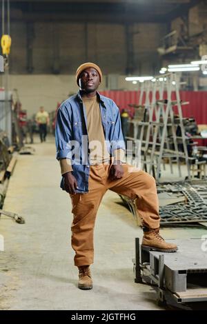 Portrait d'un jeune homme africain en vêtements de travail regardant la caméra debout dans l'atelier de l'usine Banque D'Images