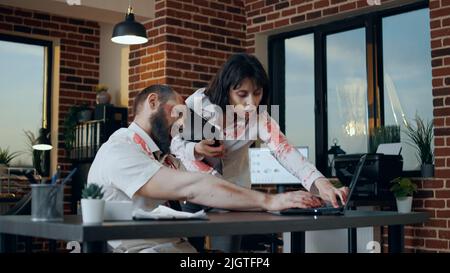 Zombie de bureau effrayant avec des blessures sanglantes dans l'espace de travail travaillant sur un ordinateur portable moderne. Monstre maléfique de cerveau-manger avec des cicatrices profondes essayant d'utiliser l'ordinateur quand la femme de marche créepy mort vient et aide. Banque D'Images