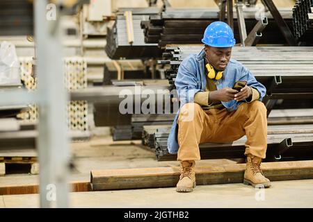 Jeune travailleur africain de hardhat défilant dans son smartphone pendant la pause à l'usine Banque D'Images