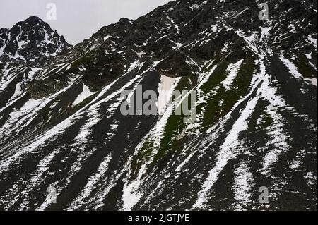 En juillet, la neige striée et le lichen vert foncé se trouvent sur des pentes de criques noires dans le canton de Graubunden, en Suisse, au-dessus du Schottensee, un arrêt tranquille sur la route sinueuse des Alpes rhétiennes reliant Davos dans la vallée de Landwasser à Susch dans la Basse-Engadine, Via le col de la Fluela à une altitude de 2 383 m ou 7 818 ft. Banque D'Images