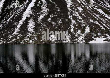 Des stries de neige couchée sur des criques noires accidentées descendant jusqu'au Schottensee se reflètent dans les eaux froides et sombres du lac, le jour de juillet gris dans le canton de Graubunden, en Suisse. Le Schottensee est un arrêt tranquille sur la route des Alpes rhétiennes reliant Davos dans la vallée de Landwasser à Susch dans la Basse-Engadine, via le col de la Fluela à une altitude de 2 383 m ou 7 818 ft. Banque D'Images