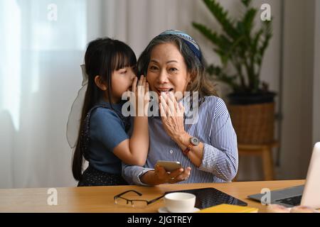 Un petit enfant mignon chuchote à l'oreille, en racontant le secret à la grand-mère tout en appréciant les loisirs ensemble à la maison Banque D'Images