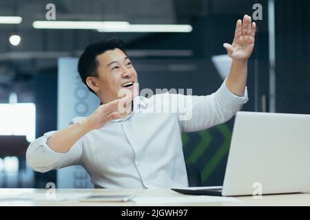 Homme d'affaires asiatique joyeux et réussi dansant au bureau pour célébrer le triomphe et la victoire assis au bureau avec un ordinateur portable Banque D'Images