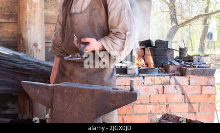 Un forgeron médiéval dans des vêtements traditionnels avec un tablier en cuir forge un fer à cheval sur une enclume. Le forgeron se tient à la forge four et hea Banque D'Images