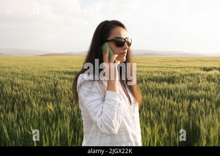 Agricultrice, agricultrice du millénaire caucasien parlant au téléphone. Femme agricole moderne ou agronome debout dans le champ de blé vert. Banque D'Images