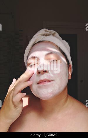 La femme Candid applique un masque d'argile, de minéraux et de sels à son visage dans la salle de bains à la maison. Soin de la peau pendant Covid-19, vraie femme, le moment de style de vie candid Banque D'Images