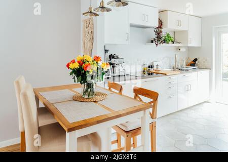 Intérieur élégant et moderne de boho, scandi d'espace ouvert cuisine blanche avec des roses jaunes et orange fleurs dans un vase sur la table de comptoir de cuisine en bois. Banque D'Images
