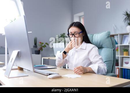 Se sentir mal au travail.Une jeune femme d'affaires asiatique, indépendant, directeur, employé est tombé malade.il est assis à la réception à l'ordinateur, essuyant le nez avec une serviette, a un nez qui coule et se sent mal. Banque D'Images