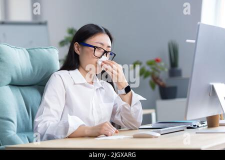 Se sentir mal au travail. Une jeune femme d'affaires asiatique est tombée malade. Il est assis au bureau de l'ordinateur dans une grande chaise, essuyant le nez avec une serviette, a un nez qui coule et se sent mal. Banque D'Images