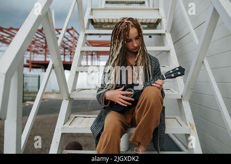 Jouer à un guitalele petite fille adorable avec des semelles assises sur un escalier en acier. Une veste grise sur un pantalon et une chemise marron assortis. Banque D'Images