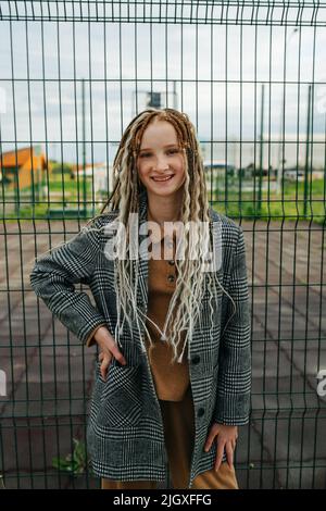 Adolescente légère avec des semelles debout devant une clôture en métal. Elle porte une veste. Portrait. Banque D'Images