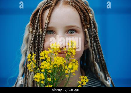 Gros plan d'une adolescente insouciante regardant l'appareil photo, tenant des fleurs jaunes. Elle a des reads. En face d'un arrière-plan flou. Banque D'Images