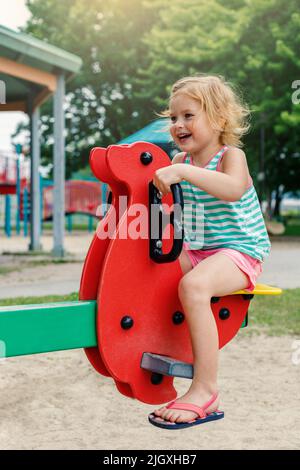 Petite fille jouant dans le parc à l'aire de jeux . Enfant souriant assis sur une balançoire de mer lors d'une journée ensoleillée d'été. Bonne enfance Banque D'Images