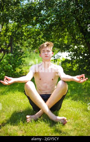 Un adolescent en short et en peluche médite sur l'herbe verte, lors d'une journée d'été ensoleillée et claire Banque D'Images