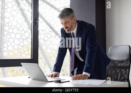 Homme d'affaires stressé d'âge moyen debout près du bureau et regardant l'écran d'ordinateur portable Banque D'Images