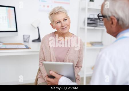 Vue sur l'épaule d'un médecin âgé utilisant un comprimé tout en montrant les résultats des tests au patient senior au rendez-vous Banque D'Images