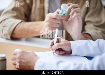 Gros plan d'un médecin masculin méconnaissable assis à une table et remplissant une ordonnance pendant que le patient lit l'étiquette du flacon de pilules Banque D'Images