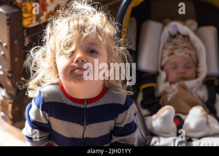 Gros plan sur une photo sélective d'un petit garçon caucasien de deux ans aux cheveux blonds ondulés et d'une sœur de nouveau-né floue dans un siège de bébé en arrière-plan. Banque D'Images