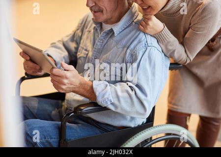 Gros plan d'un couple d'âge positif regardant une vidéo sur une tablette numérique : femme souriante se penchant sur un fauteuil roulant de son mari Banque D'Images
