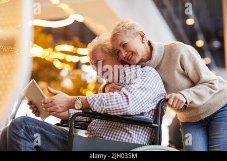Heureux aimant belle femme mûre embrassant la personne handicapée mari en fauteuil roulant pendant qu'il regarde la vidéo sur tablette Banque D'Images