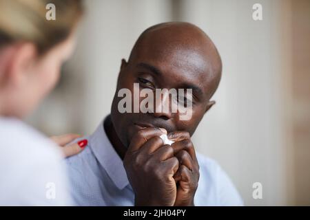 Gros plan du jeune homme noir bald déprimé avec des yeux tristes tenant la serviette dans les mains et pleurant tout en parlant au thérapeute à la séance de thérapie Banque D'Images