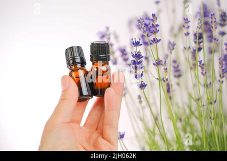 Huile essentielle de lavande en bouteilles de verre dans une main femelle et fleurs sur fond blanc. Essence avec parfum de lavande. Aromathérapie et massage Banque D'Images