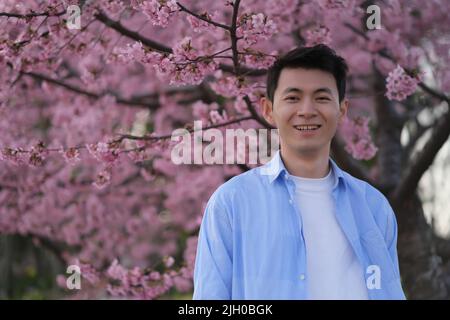 Souriant beau jeune homme asiatique, flou rose kawazu-zakura fleur de fond d'arbre Banque D'Images