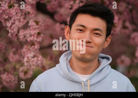 Adorable adorable jeune homme asiatique faisant le visage, regardant l'appareil photo. Flou rose sakura fleurs fond Banque D'Images