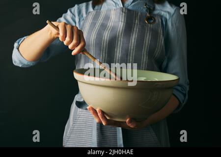 Croiser la femme anonyme cuire en tablier en mélangeant les ingrédients dans un bol avec une spatule en bois tout en préparant la cuisson sur fond noir Banque D'Images