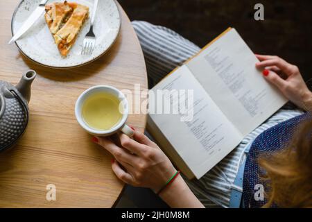 Mains de femme avec manucure rouge tenant livre ouvert, tasse de thé et tarte sur table en bois clair, vue du dessus. Banque D'Images