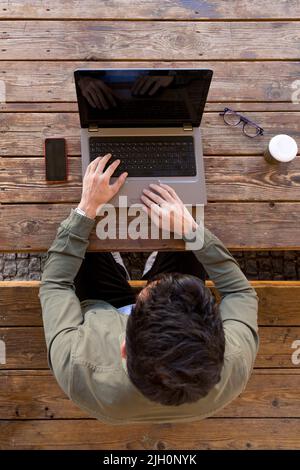 Vue de dessus d'un homme travaillant avec un ordinateur portable assis sur une table en bois d'une terrasse à l'extérieur Banque D'Images