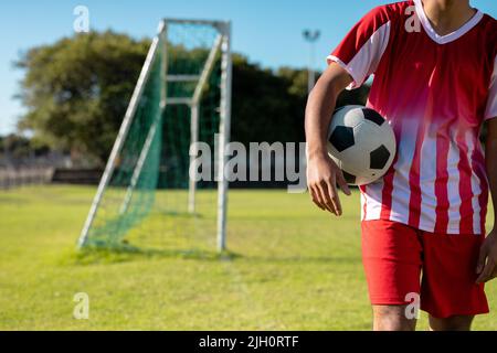 Section médiane d'un joueur masculin caucasien portant un maillot rouge avec une balle de football debout sur des terres herbeuses Banque D'Images