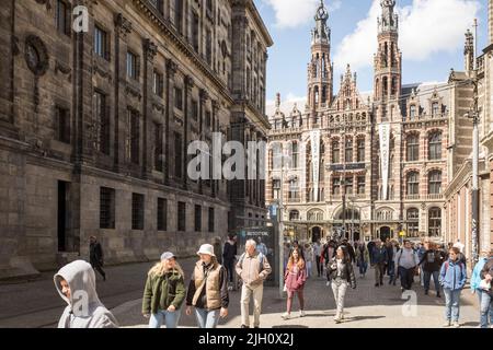 Les gens marchent près du Palais Royal sur la place Dam sous le soleil du printemps avec le centre commercial néo-gothique Magna Plaza à Amsterdam, pays-Bas. Banque D'Images