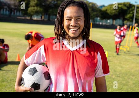 Portrait d'un joueur biracial masculin avec des dreadlocks tenant le ballon de football et riant à l'aire de jeux Banque D'Images