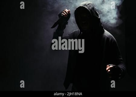 Silhouette de bandit afro-américain à capuche tenant le couteau sur fond noir avec de la fumée Banque D'Images