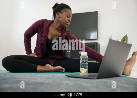 Gros plan de la jeune femme africaine noire après un cours de fitness en ligne sur ordinateur portable, les yeux étirés fermés, l'exercice sur le tapis de yoga dans le salon moderne à Banque D'Images