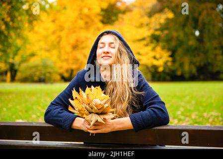 jolie femme blonde portant un long sweat à capuche avec capuche sur la tête est assise sur un banc et tenant des feuilles d'érable jaune, appréciant à l'extérieur dans le parc de la ville Banque D'Images