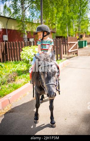 Belle petite fille de deux ans équitation poney cheval dans grand casque de sécurité jockey posant à l'extérieur sur la campagne Banque D'Images