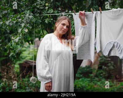 femme caucasienne d'âge moyen en robe blanche faisant des devoirs, pendant des vêtements sur la corde à linge dans la rue dans la cour de la maison de village, concept de jour de blanchisserie Banque D'Images